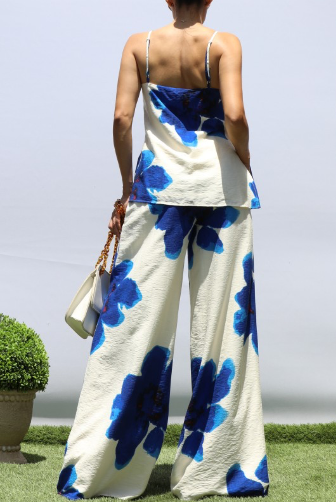 A woman shown from the back wearing a matching two-piece outfit with a white base and bold blue floral print, standing outdoors on grass with a pale sky and potted plants in the background.