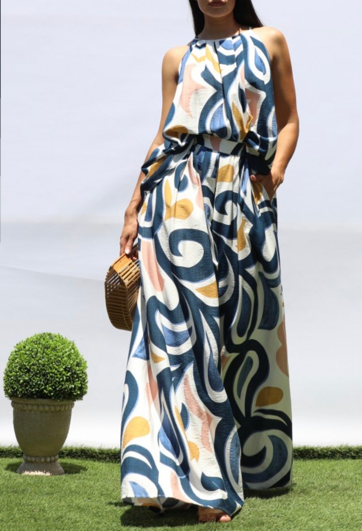 A woman wearing a sleeveless, high-neck, floor-length maxi dress with an abstract wavy pattern in navy blue, beige, and mustard yellow. She is standing on artificial green grass against a white background, holding a small woven brown handbag.
