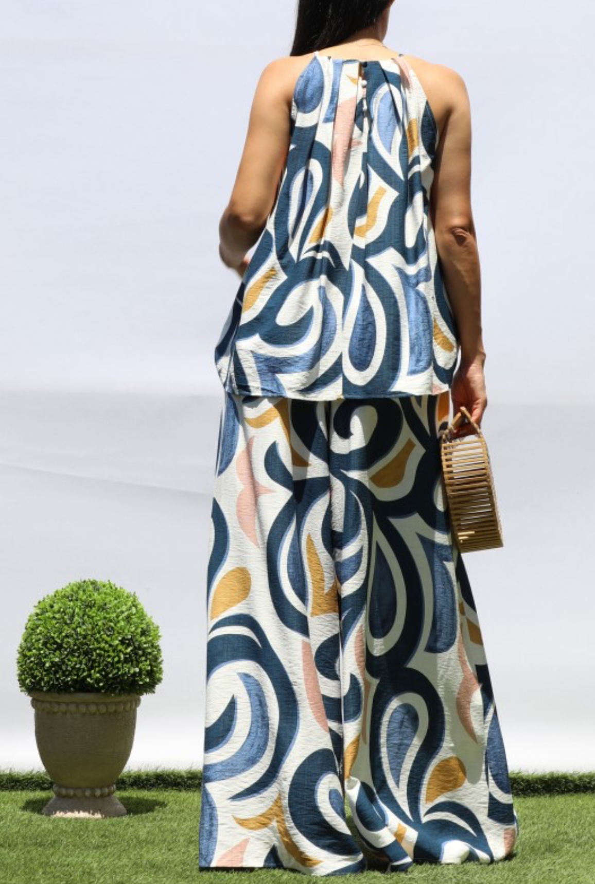 A woman viewed from behind wearing a sleeveless, floor-length maxi dress with a bold abstract swirl pattern in navy blue, beige, and mustard yellow on a white background. She is standing outdoors on a green lawn next to a potted topiary tree.