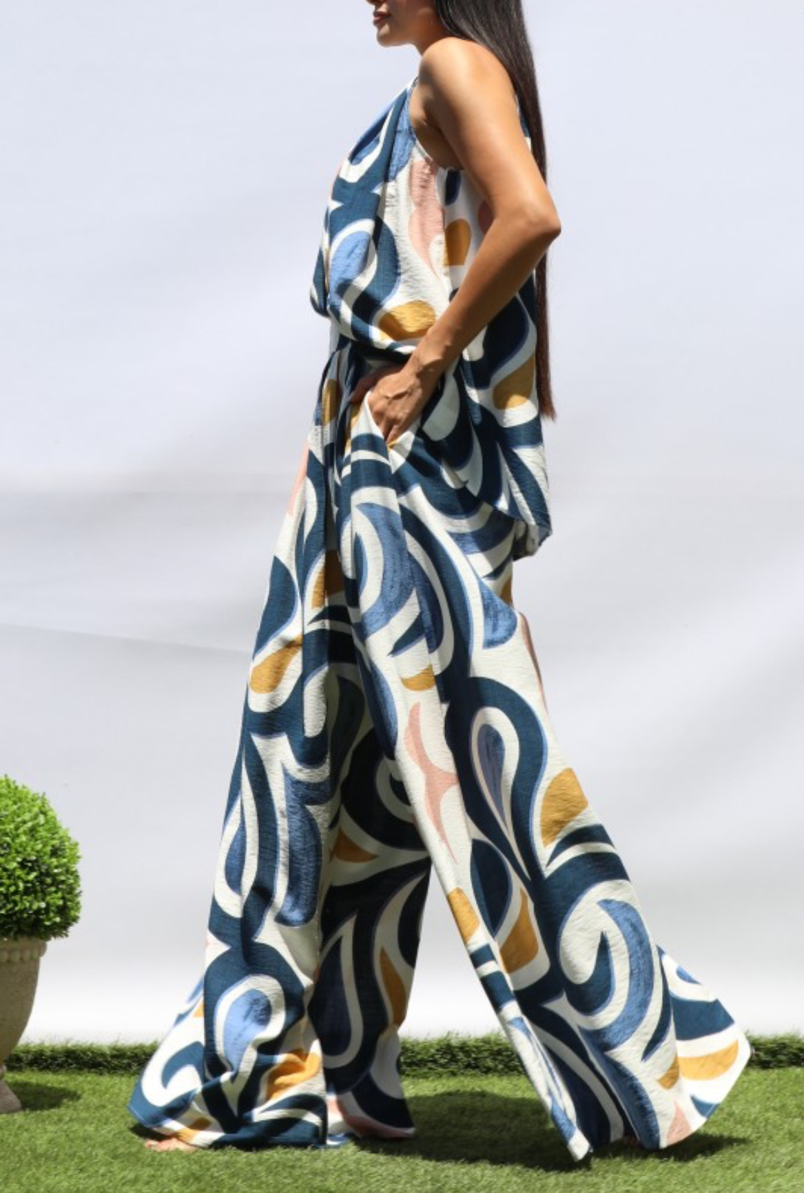 A woman modeling a sleeveless, wide-leg jumpsuit with an abstract swirl pattern in navy blue, mustard yellow, and white, set against a white studio backdrop with a hint of green grass at the bottom.