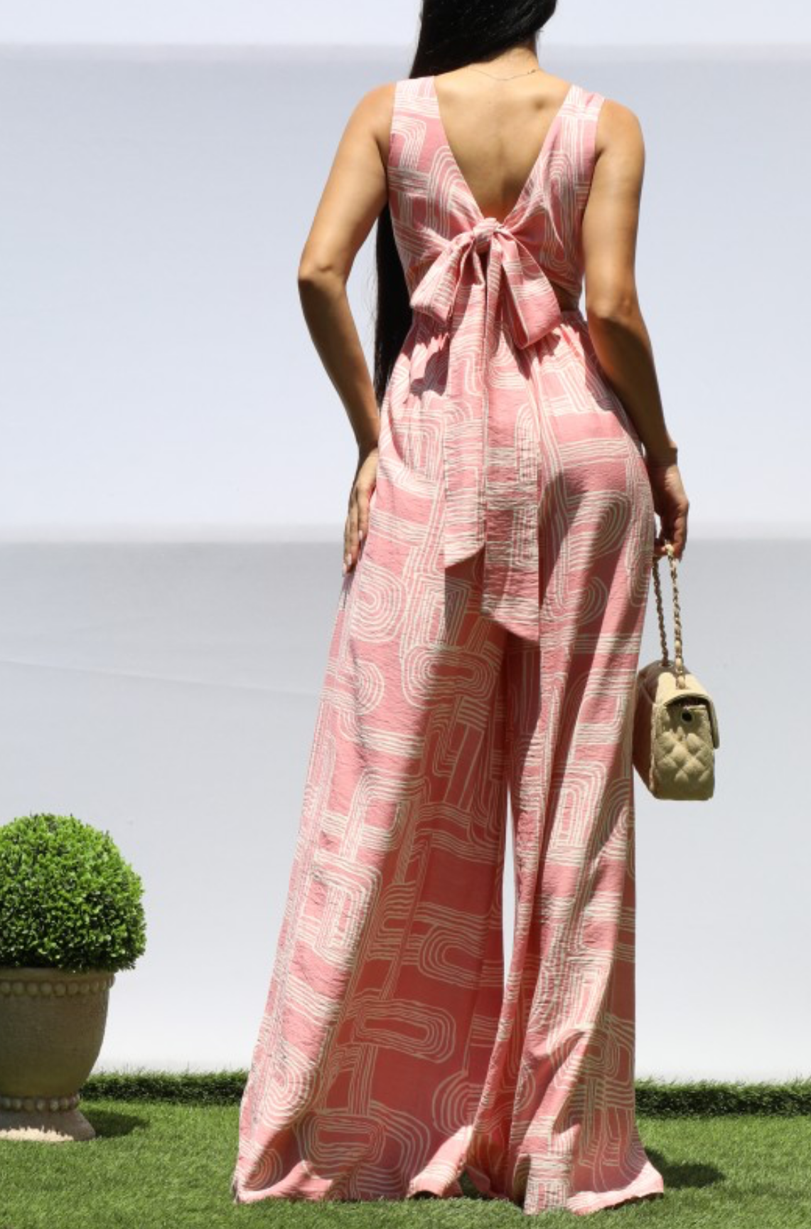A woman stands outdoors on grass wearing a sleeveless pink patterned jumpsuit with a deep V-back and large tie detail at the back. She is shown from behind, holding a small quilted handbag.