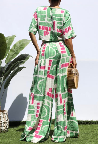 A woman shown from the back wearing a matching green, pink, and white geometric print two-piece set with short sleeves and wide-leg pants, standing outdoors on grass against a white wall and holding a woven straw bag.