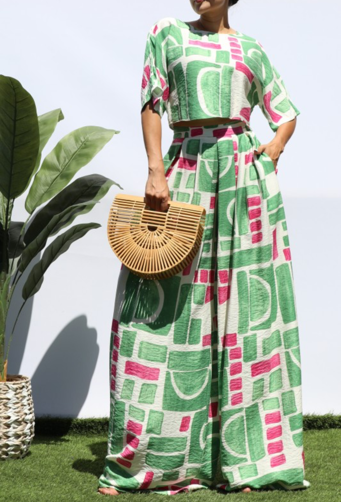 A woman modeling a matching two-piece set featuring a green, pink, and white geometric pattern, holding a round woven straw bag.