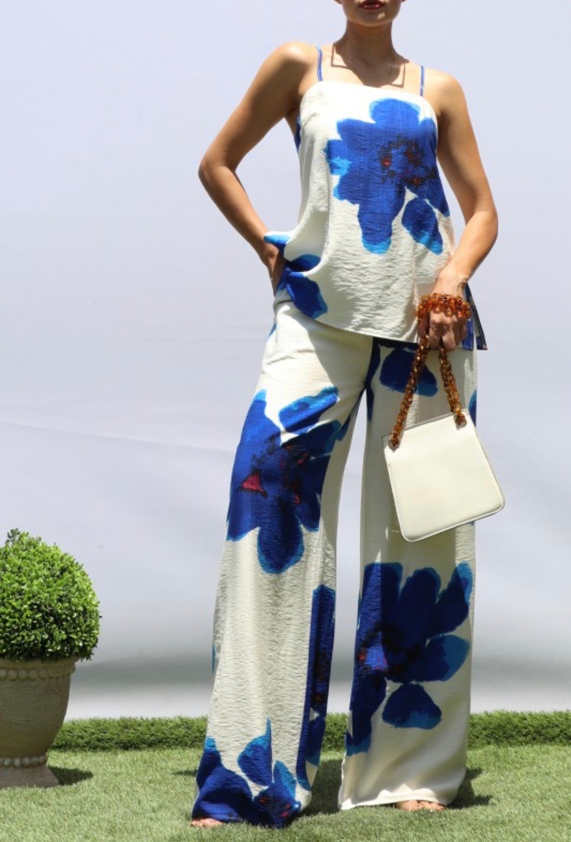 A woman modeling a sleeveless white two-piece set with large blue floral prints, standing on artificial grass against a light gray backdrop and holding a white handbag.