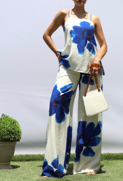 A woman modeling a sleeveless white two-piece set with large blue floral prints, standing on artificial grass against a light gray backdrop and holding a white handbag.