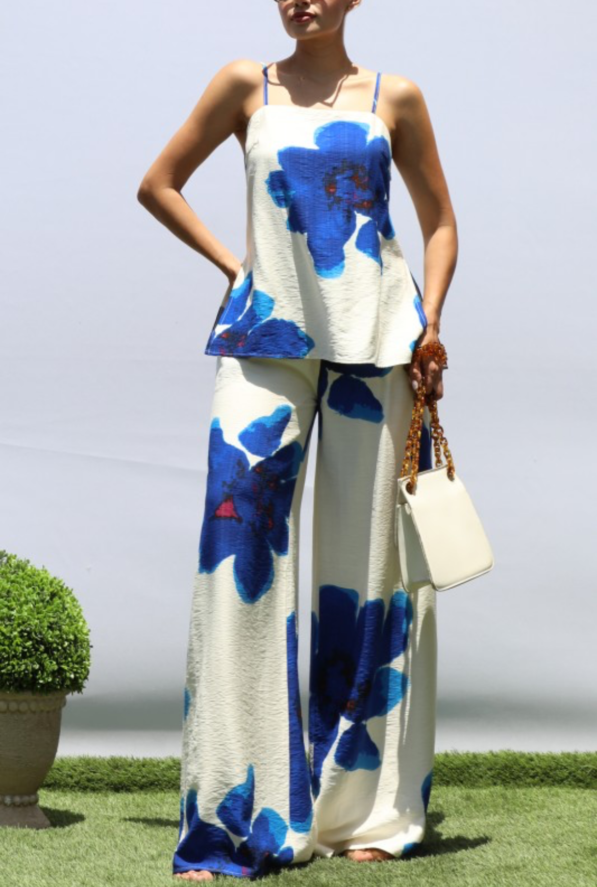A woman modeling a sleeveless white two-piece set with large blue floral prints, standing outdoors on grass against a pale sky. She is holding a small white handbag.