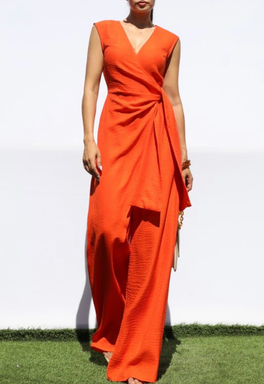 A woman wearing a sleeveless, wrap-style maxi dress in a vibrant orange color, standing outdoors on a patch of green grass against a white background.