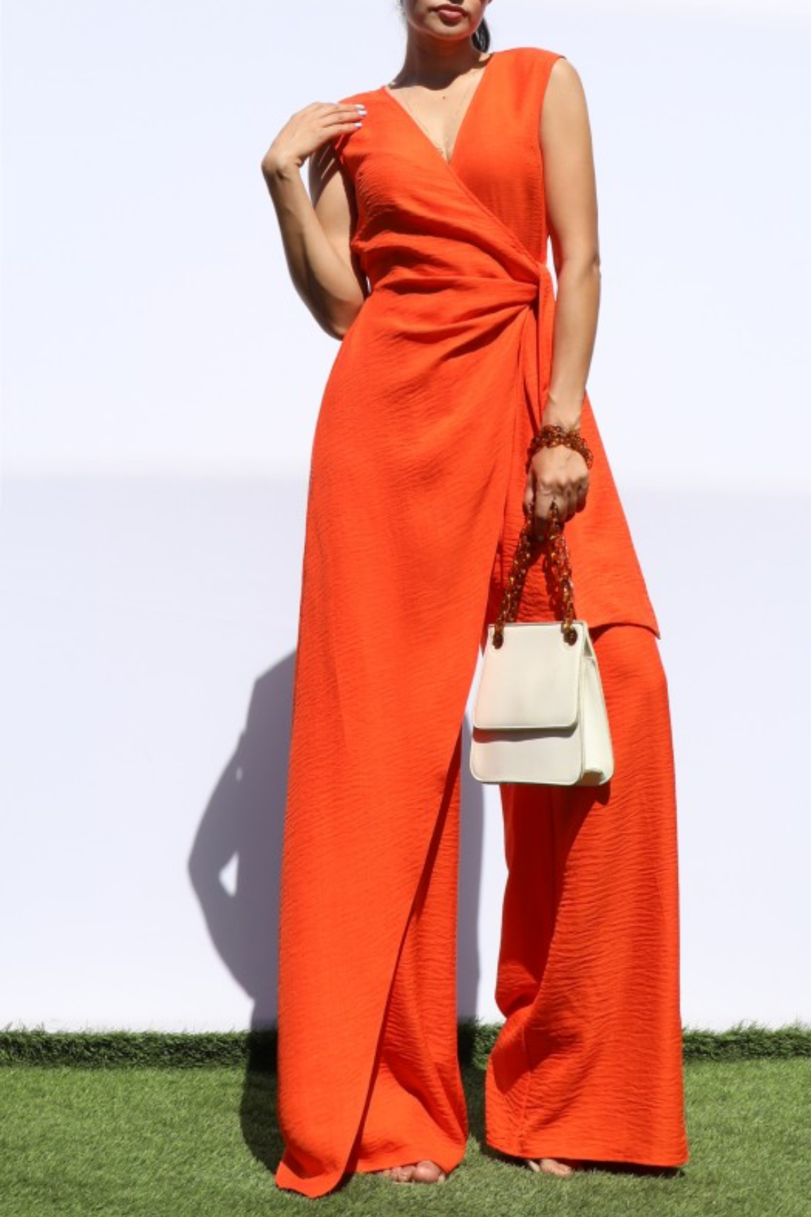 A woman modeling a bright orange sleeveless wrap-style jumpsuit with wide legs, standing outdoors against a white wall and green grass, holding a small cream handbag.