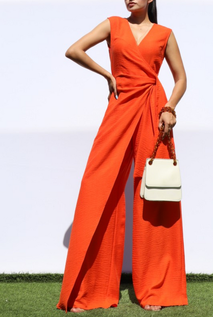 A woman modeling a bright orange sleeveless wide-leg jumpsuit with a V-neck and gathered waist detail, holding a small white handbag, standing outdoors on grass against a white wall.