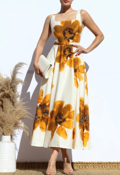 A woman models a sleeveless midi dress with a white base and large mustard yellow floral print. She is holding a white clutch and wearing beige high-heeled sandals, standing against a white wall with dried pampas grass in a white vase to her side.