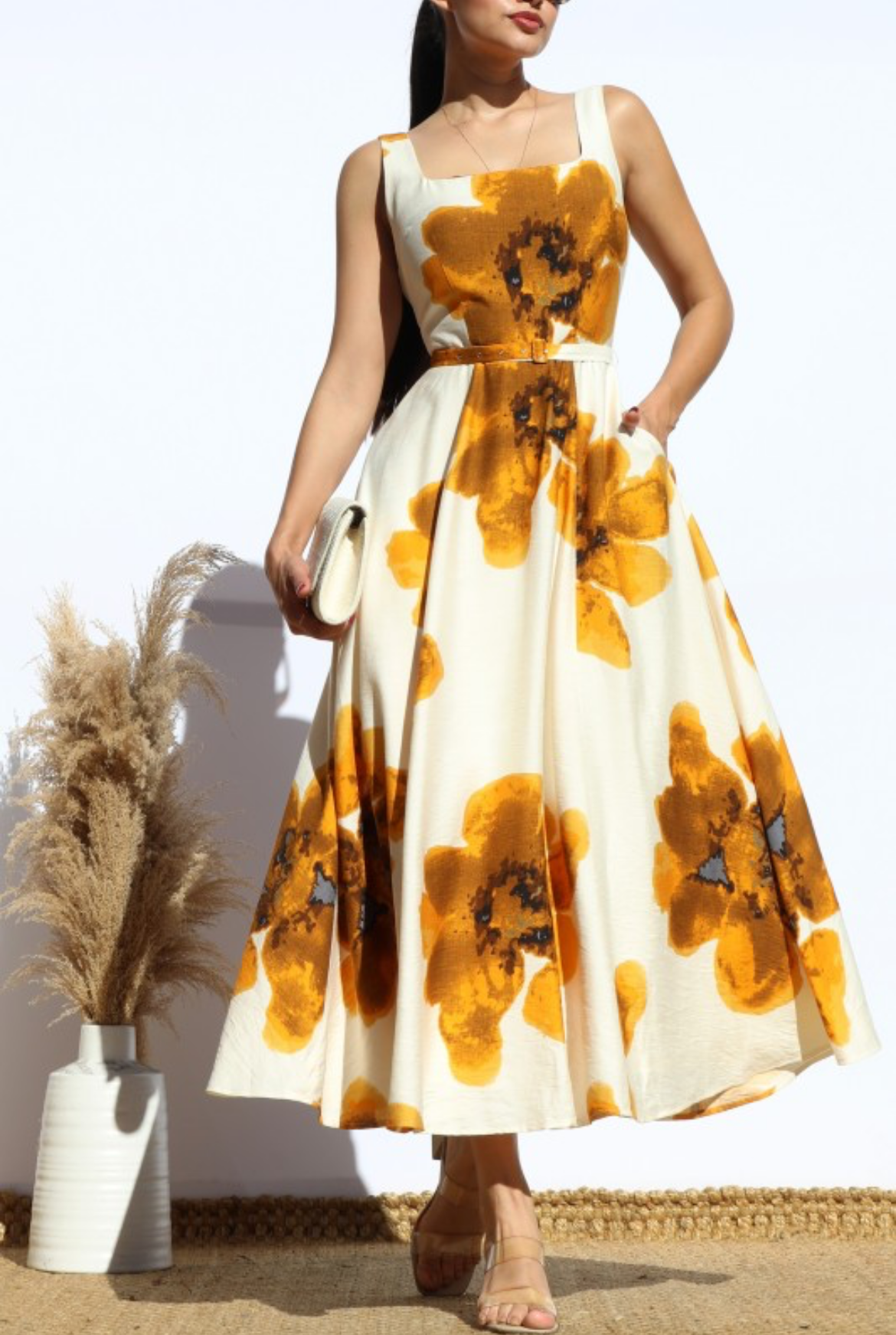 A woman wearing a sleeveless midi dress with a large yellow and orange floral print on a cream background, standing in a sunlit indoor setting with pampas grass in a vase.
