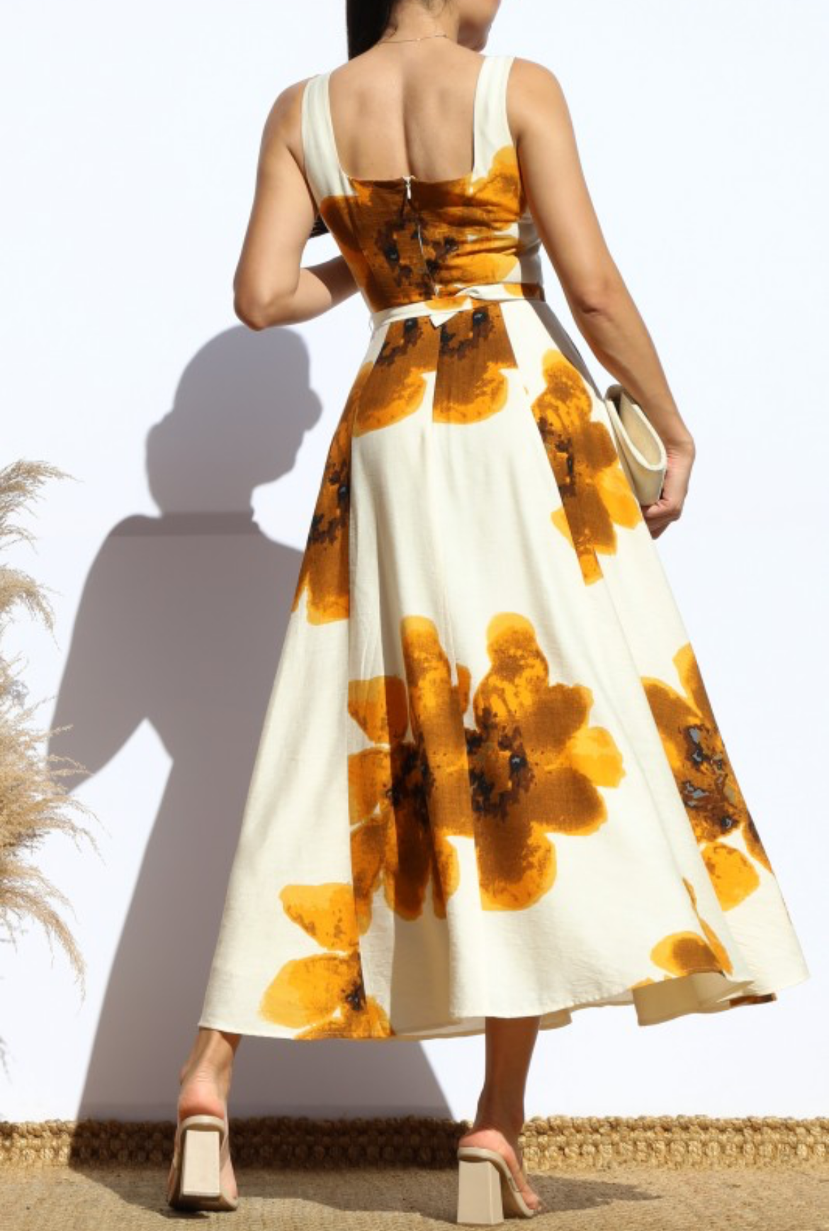 A woman wearing a sleeveless midi dress with a white base and large mustard yellow floral print, shown from the back against a white wall.