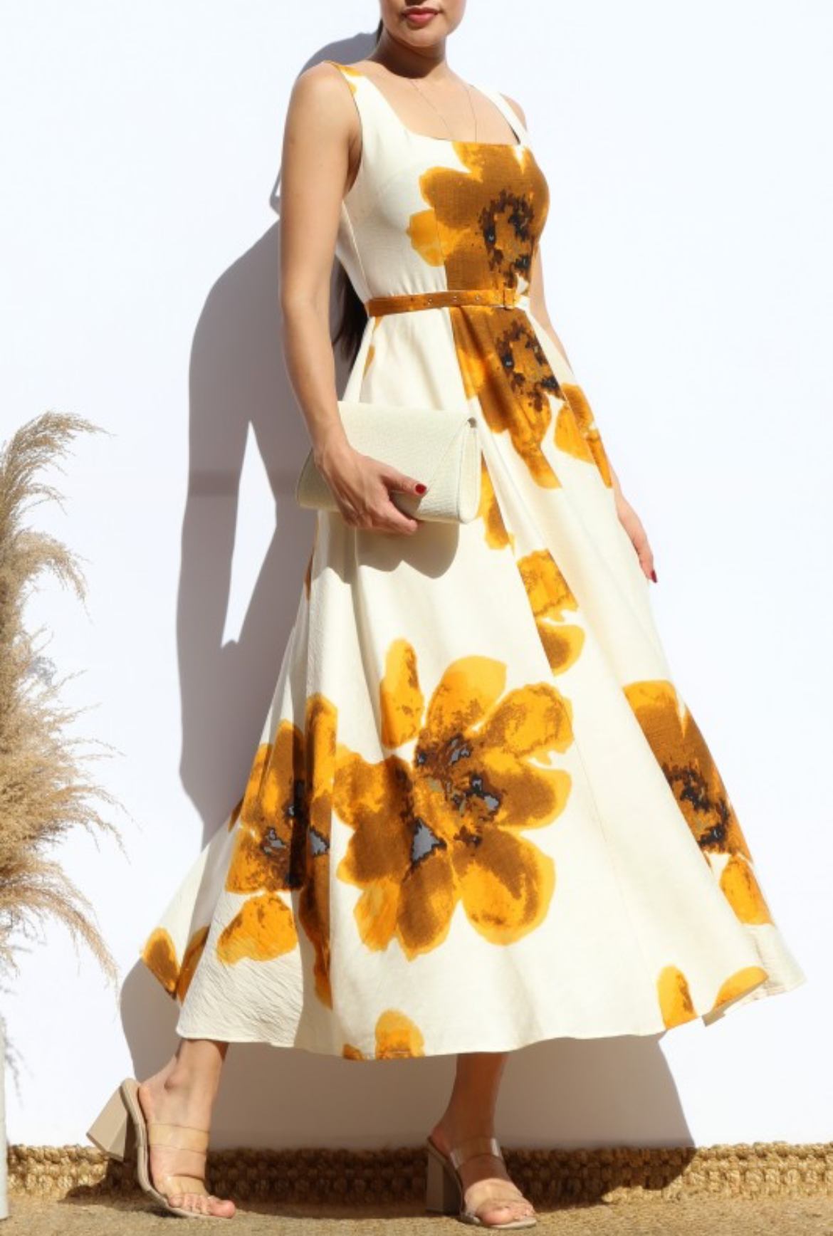 A woman models a sleeveless midi dress with a white base and large mustard yellow floral print, styled with a matching belt and holding a small white clutch.