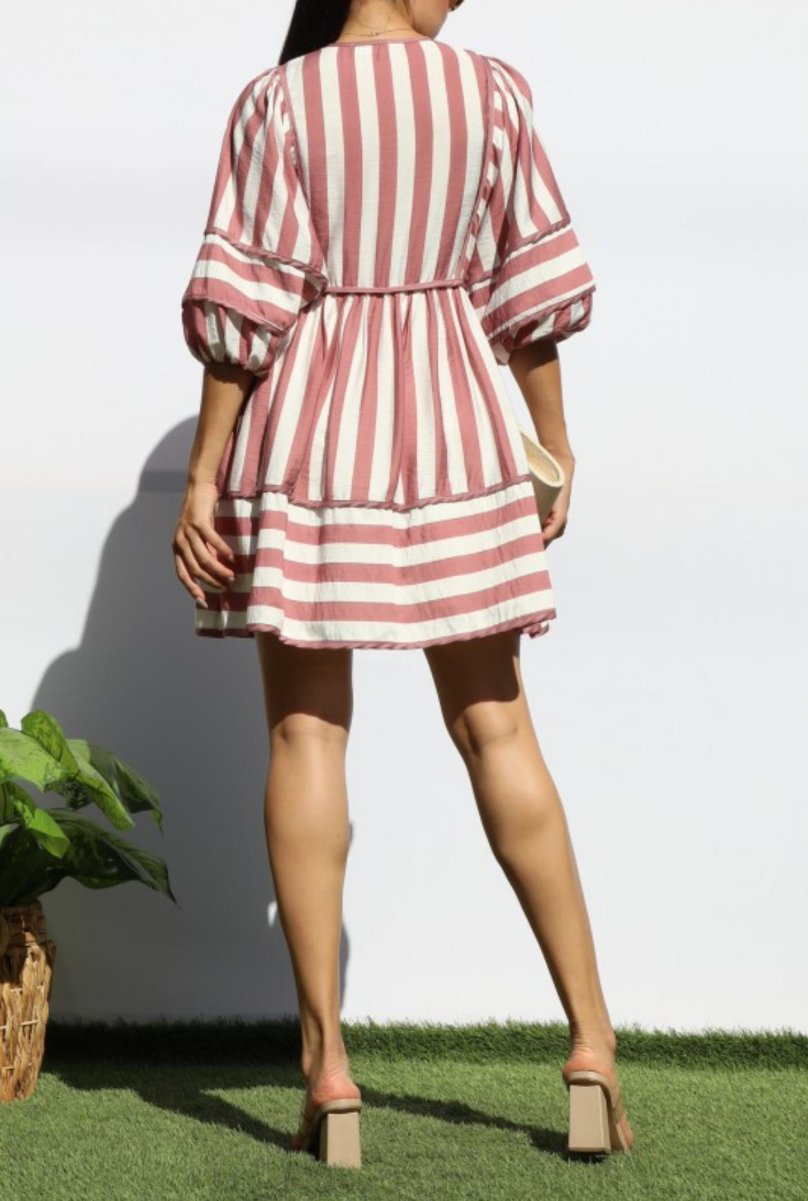 A woman shown from the back wearing a short puff-sleeve dress with vertical pink and white stripes, standing on artificial grass against a white wall.