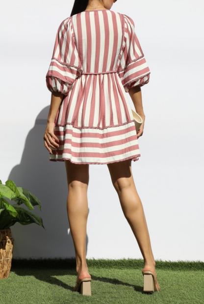 A woman shown from the back wearing a short puff-sleeve dress with vertical pink and white stripes, standing on artificial grass against a white wall.