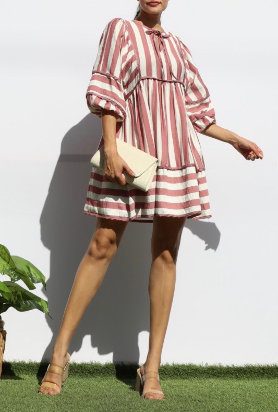 A woman models a short puff-sleeve dress with vertical dusty pink and white stripes, holding a cream clutch bag against a white wall.