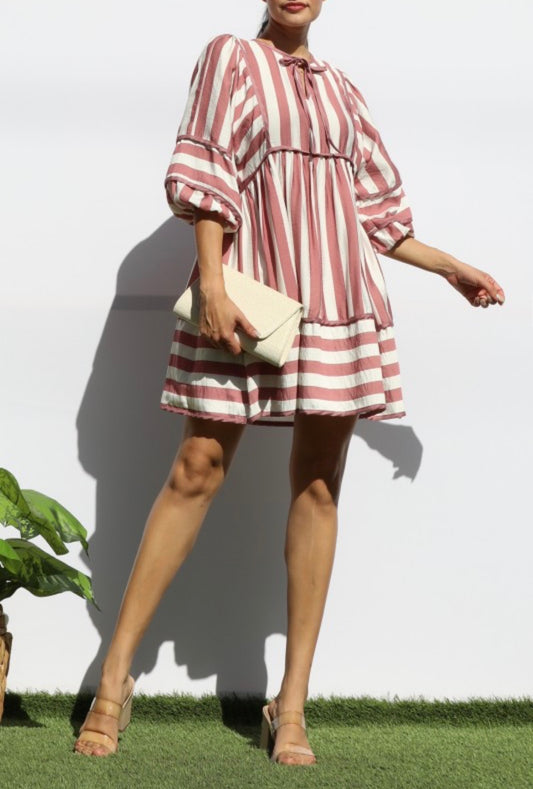 A woman models a short puff-sleeve dress with vertical dusty pink and white stripes, holding a cream clutch bag against a white wall.