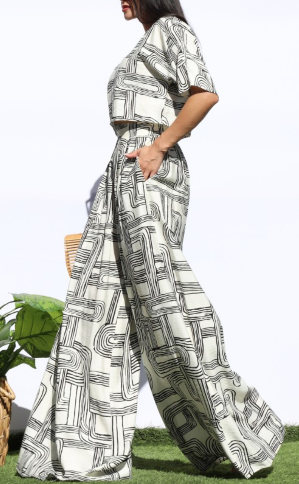 A woman wearing a matching short-sleeve top and wide-leg trousers set in an abstract black and white geometric pattern, standing outdoors on grass against a white background.