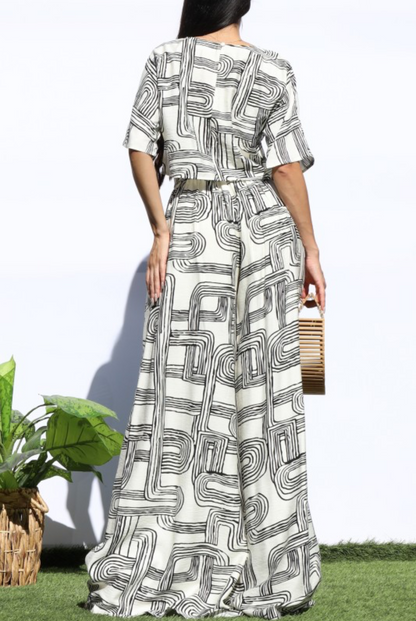A woman shown from the back wearing a long white dress with a black geometric line pattern, standing outdoors against a white wall with grass and a potted plant at the side.