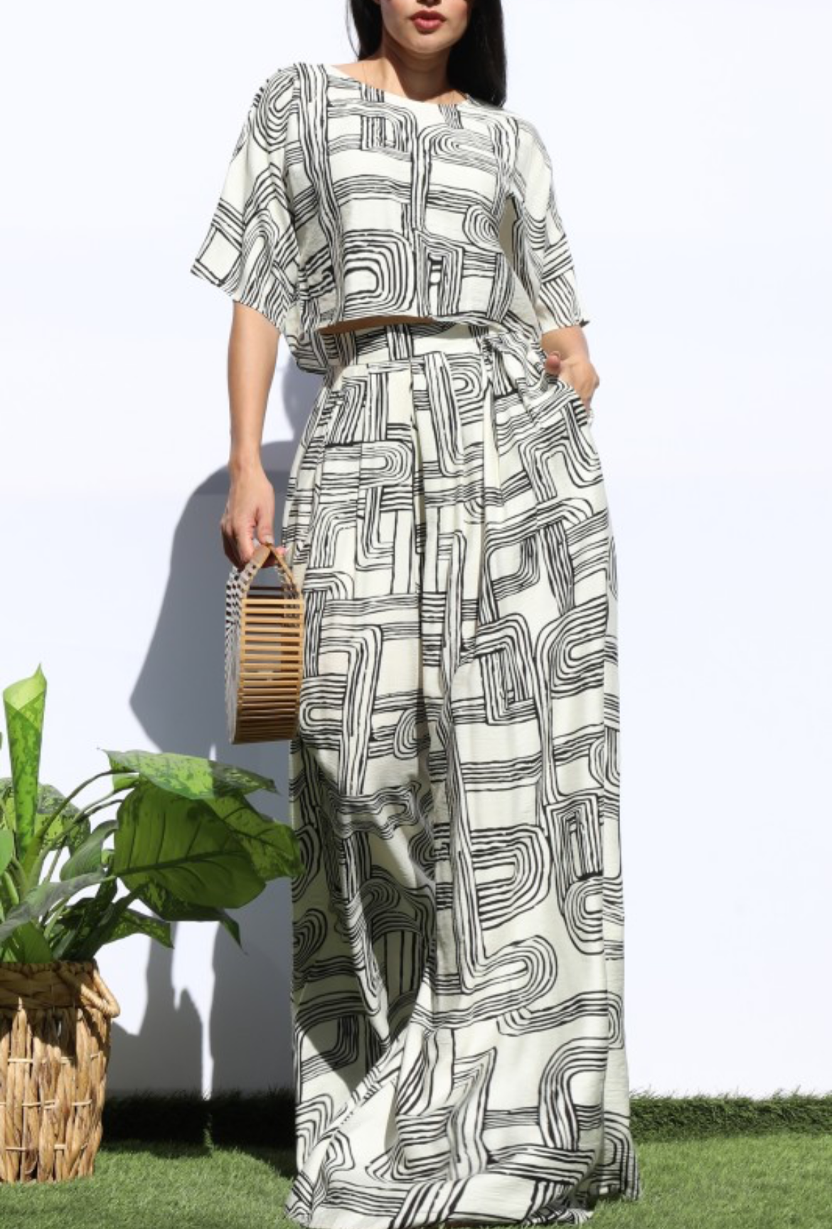 A woman modeling a matching two-piece set with an abstract black and gray geometric pattern on a light beige background, standing outdoors against a white wall with green grass and a potted plant at her side.
