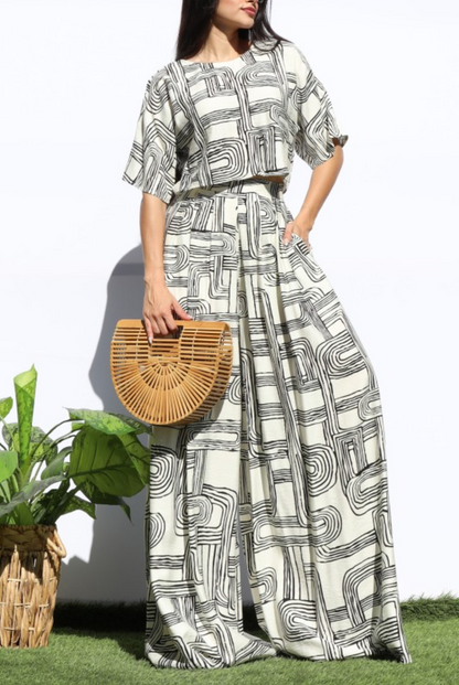 A woman models a matching two-piece set featuring a black and white abstract geometric pattern, holding a round woven straw handbag. She is standing outdoors on a patch of green grass against a white wall.