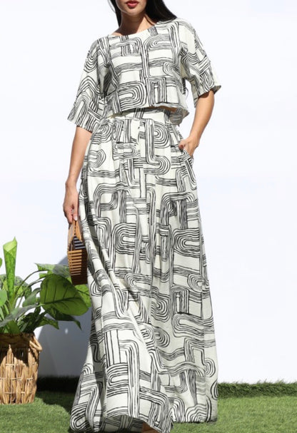 A woman modeling a matching two-piece set consisting of a short-sleeve crop top and a long maxi skirt, both featuring a black and white abstract geometric maze-like pattern. She is standing outdoors on a patch of green grass against a white wall, holding a small woven handbag.